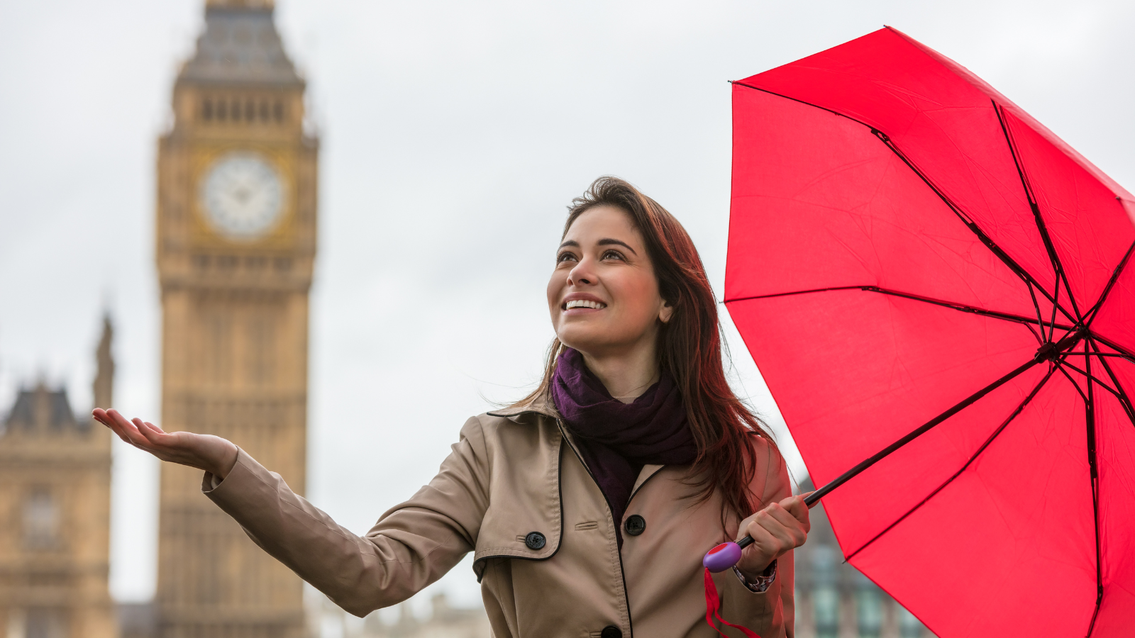 A young woman holding a red umbrella and checking for rain with Big Ben in the background.